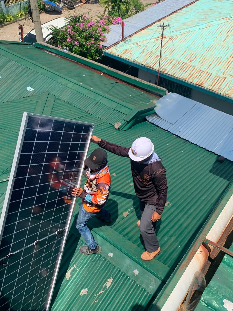 Two SolarWave installers carrying a solar panel across a green GI-sheet roof