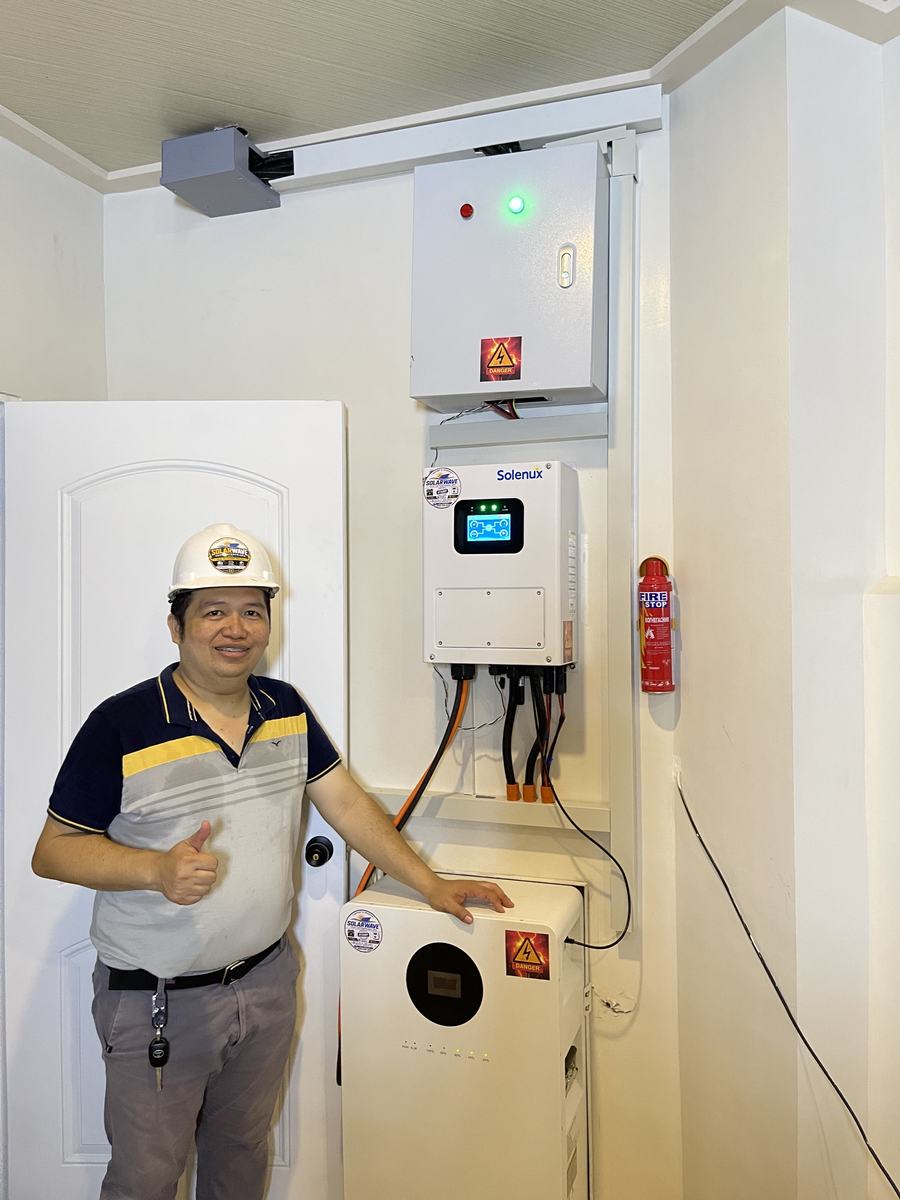 SolarWave installer in a hard hat presenting a finished wall-mounted inverter and battery installation
